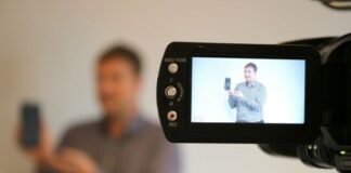 Mann steht vor Videokamera und präsentiert ein Smartphone in seiner Hand. Foto: Verbraucherzentrale NRW.
