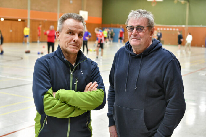 Zwei Trainer in Sporthalle mit Sportlern im Hintergrund.