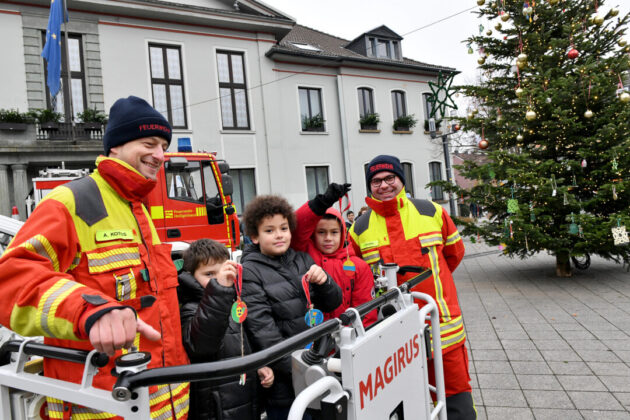 Feuerwehrleute und Kinder vor Weihnachtsbaum mit Feuerwehrauto.