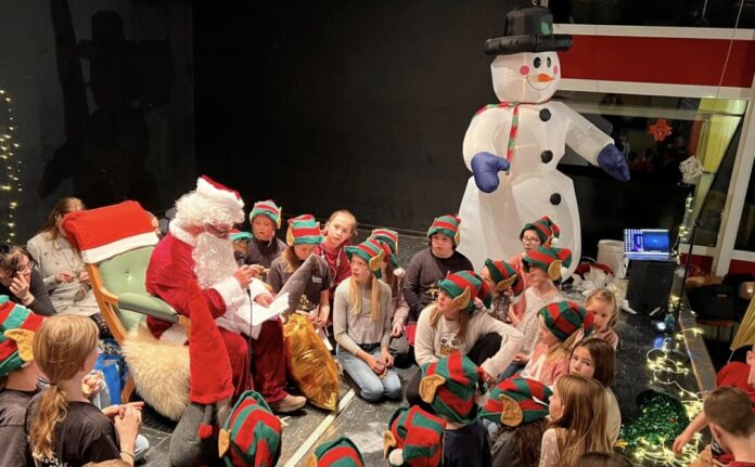 Weihnachtsmann liest Kindern in Elfenmützen vor.