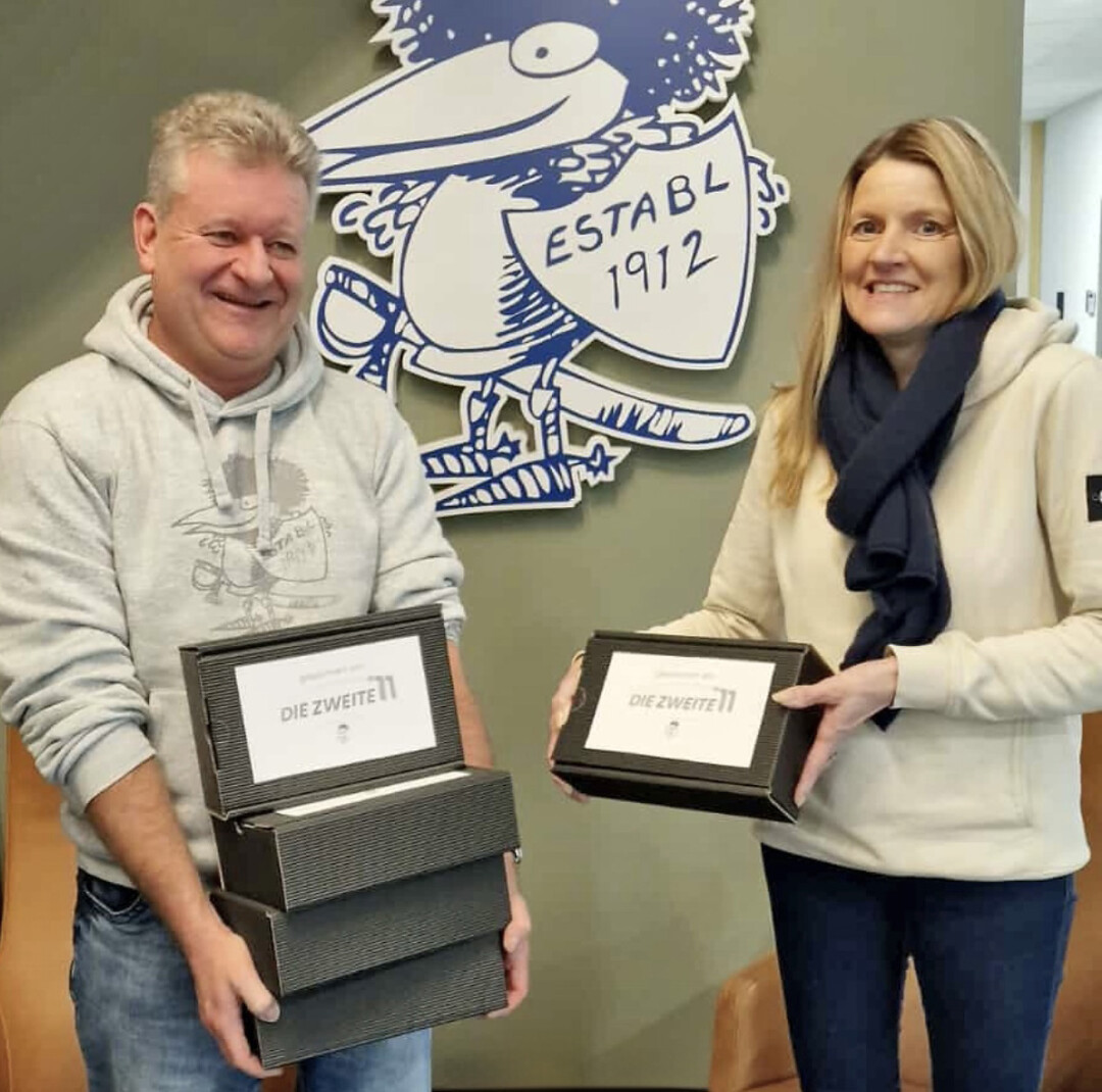 Zwei Personen präsentieren Boxen vor Storch-Maskottchen.