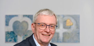 Lächelnder älterer Herr mit Brille, Anzug und Gemälde.