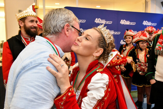 Karnevalskönigin küsst Mann auf Wange, frohe Leute im Hintergrund.