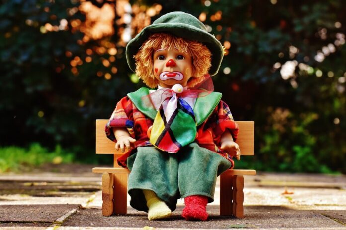 Bunter Clown-Puppe sitzt auf kleiner Holzbank.