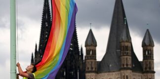 Ein Mann hisst eine Regenbogenfahne vor der Kulisse des Kölner Doms und der romanischen Kirche Groß St. Martin - eben das soll bei der Eröffnung eines erzbischöflichen Bildungszentrums nicht erwünscht gewesen sein (Archivbild). 