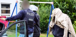 Ein Polizist führt zwei Personen in Herdecke, die ihre Gesichter verdecken, zu einem Auto. 