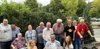 Leon Jakubeit (untere Reihe, 2.v.r.), Auszubildender bei der Sparda-Bank West, überreicht einen symbolischen Scheck an das Team des Franz Sales Haus Essen sowie einige Klientinnen und Klienten. Foto: Sparda-Bank West