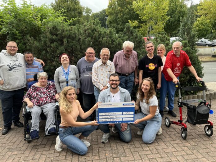 Leon Jakubeit (untere Reihe, 2.v.r.), Auszubildender bei der Sparda-Bank West, überreicht einen symbolischen Scheck an das Team des Franz Sales Haus Essen sowie einige Klientinnen und Klienten. Foto: Sparda-Bank West