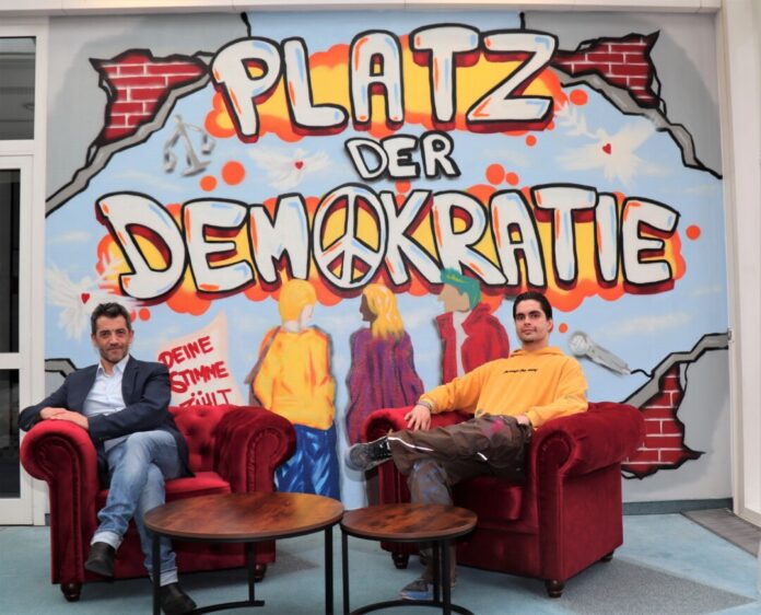 2025-12-04-Platz der Demokratie Bitte Platz nehmen: Ahmet Özdemir (links), Leiter des Medienzentrums, und Graffiti-Künstler Mel Ritschel haben die Sitzecke vor der neu gestalteten Wand im 2. Obergeschoss schon mal getestet. Foto: Stadt Ratingen