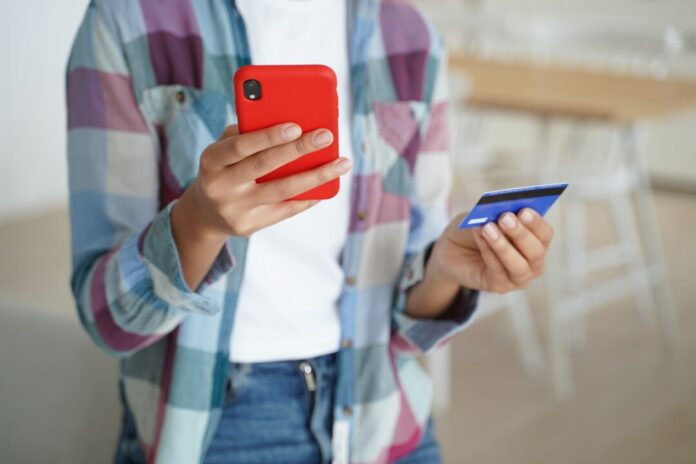 Female holding credit card, phone, uses online banking services, mobile bank apps, paying purchases Die Verbraucherzentrale NRW rät, bei Neobanken auf Extra-Kosten und Einschränkungen zu achten. Foto: VZ NRW/adpic